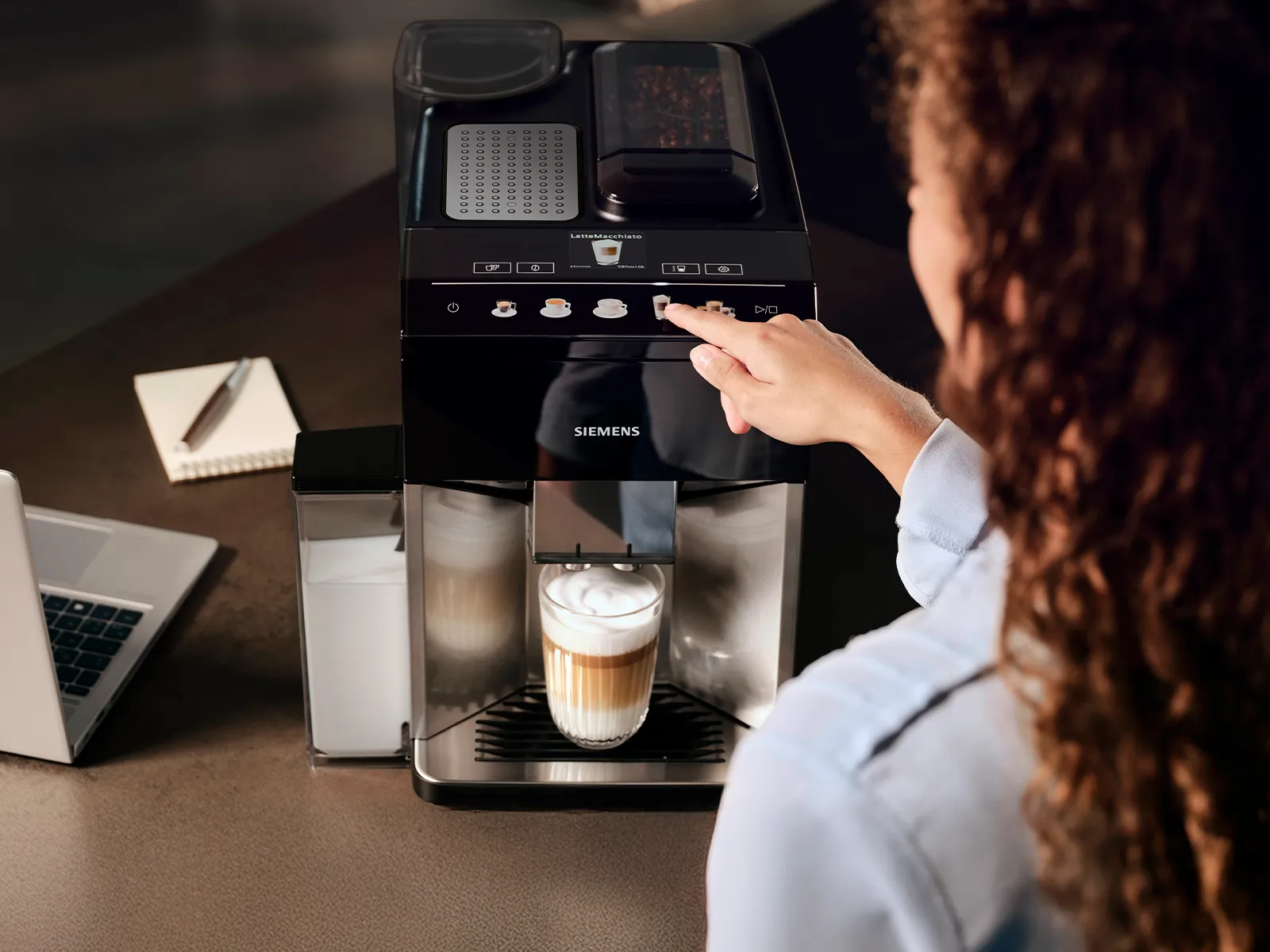 Eine Frau bereitet mit einer modernen Kaffeemaschine einen Cappuccino zu; daneben liegen ein Laptop und ein Notizblock.