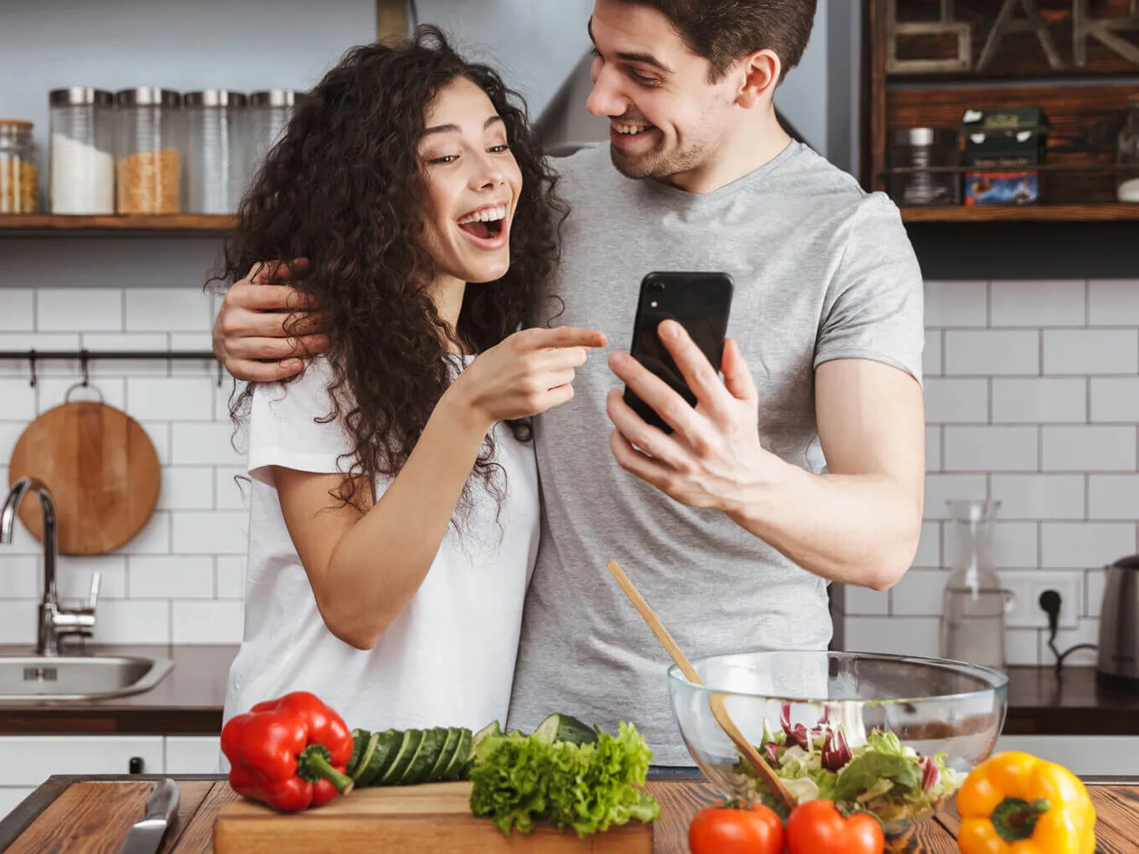 Smiling couple in a kitchen looking at a smartphone while preparing fresh vegetables and a salad.