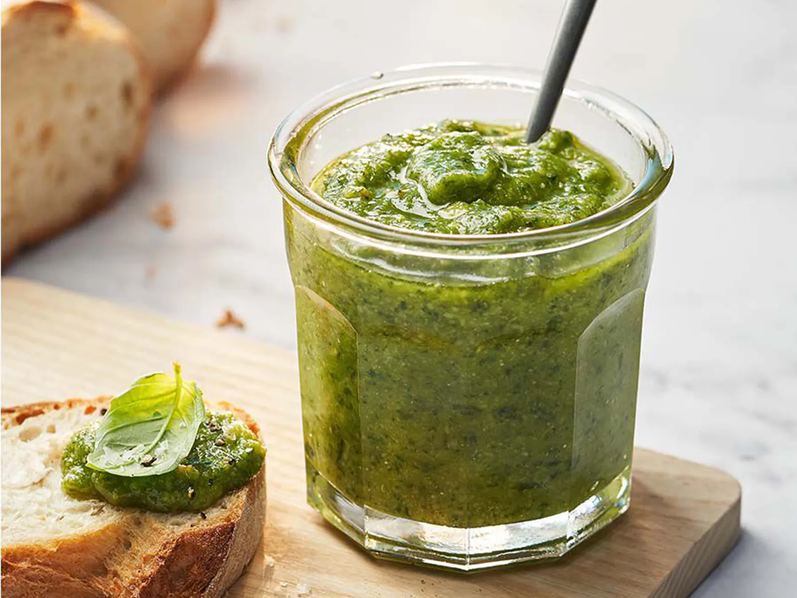 Glass jar filled with green pesto sauce, with a spoon inside, served with bread slices on a wooden board.