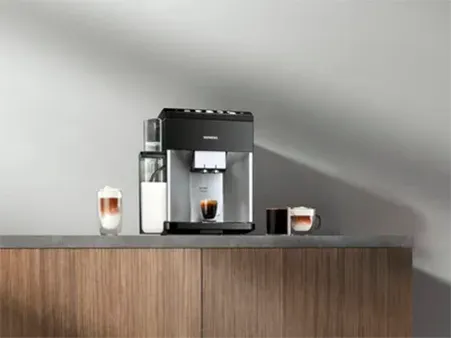A black coffee machine on a countertop, with cups of coffee and milk froth beside it, in a kitchen setting.