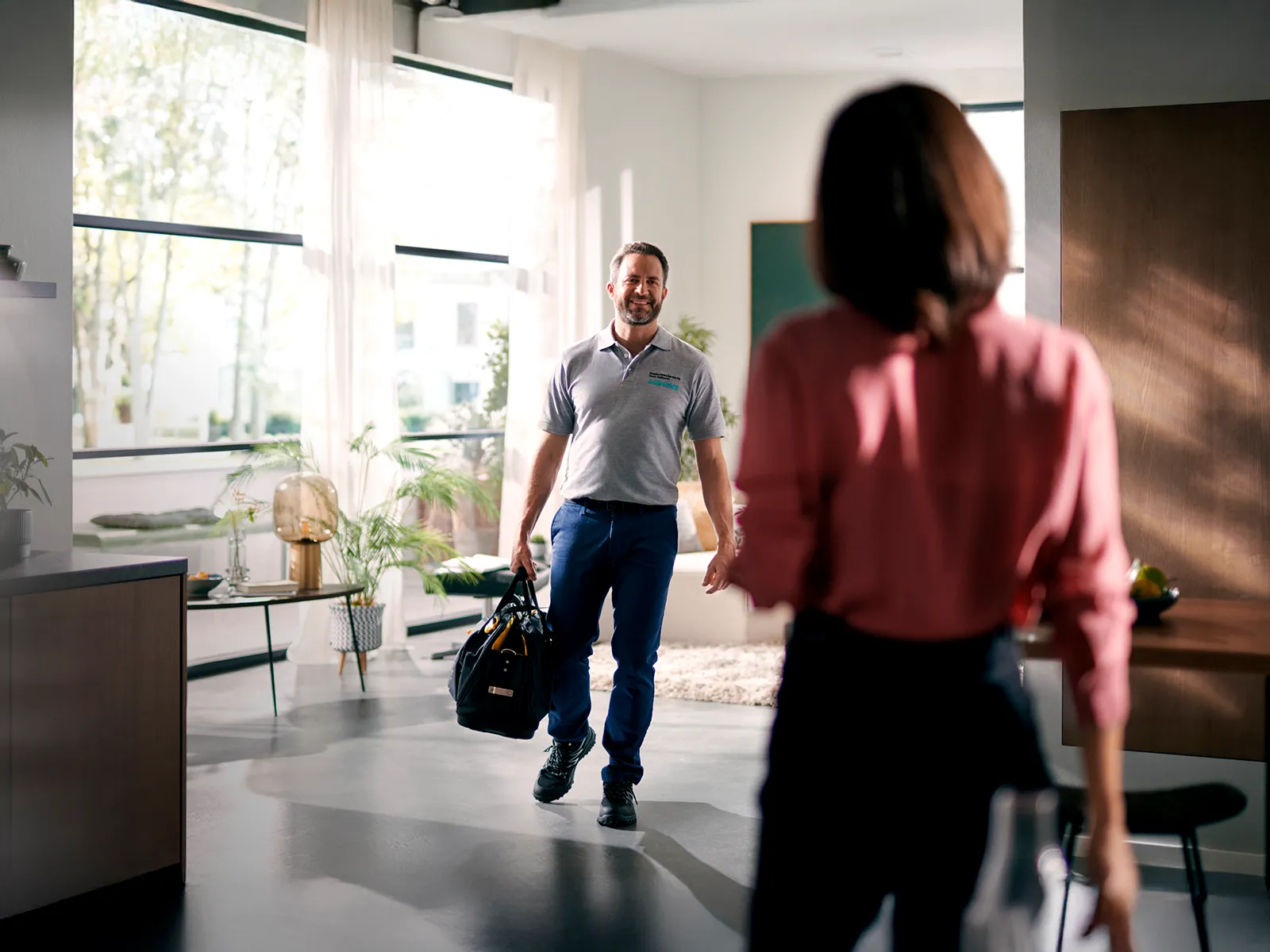 Man in a grey uniform and blue trousers, carrying a tool bag, entering a bright room while a woman, seen from behind, greets him.