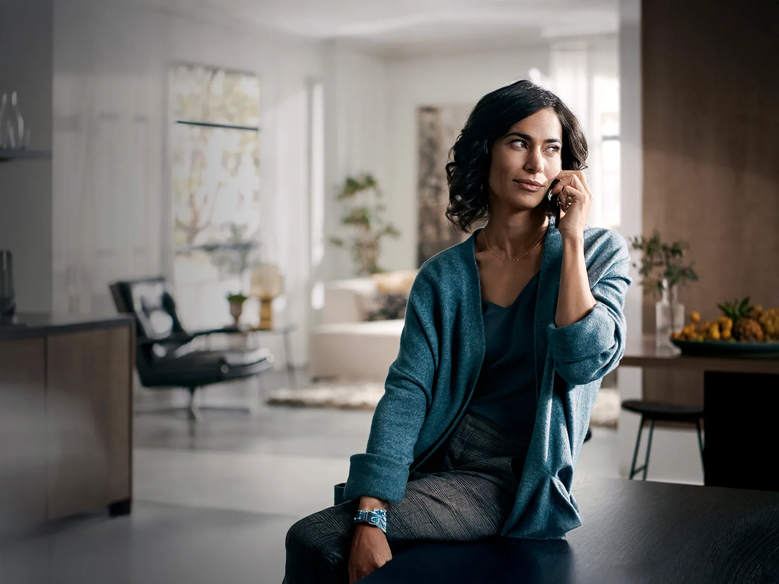 Dark-haired woman with curly hair, wearing a blue cardigan, talking on the phone in a bright, modern space with minimalist furniture.