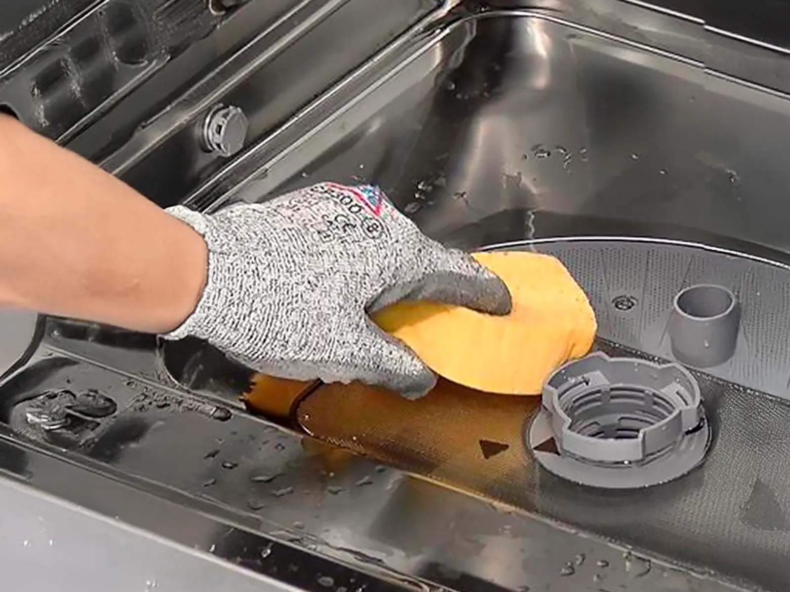 Gloved hand cleaning the filter inside a stainless steel dishwasher with a sponge.