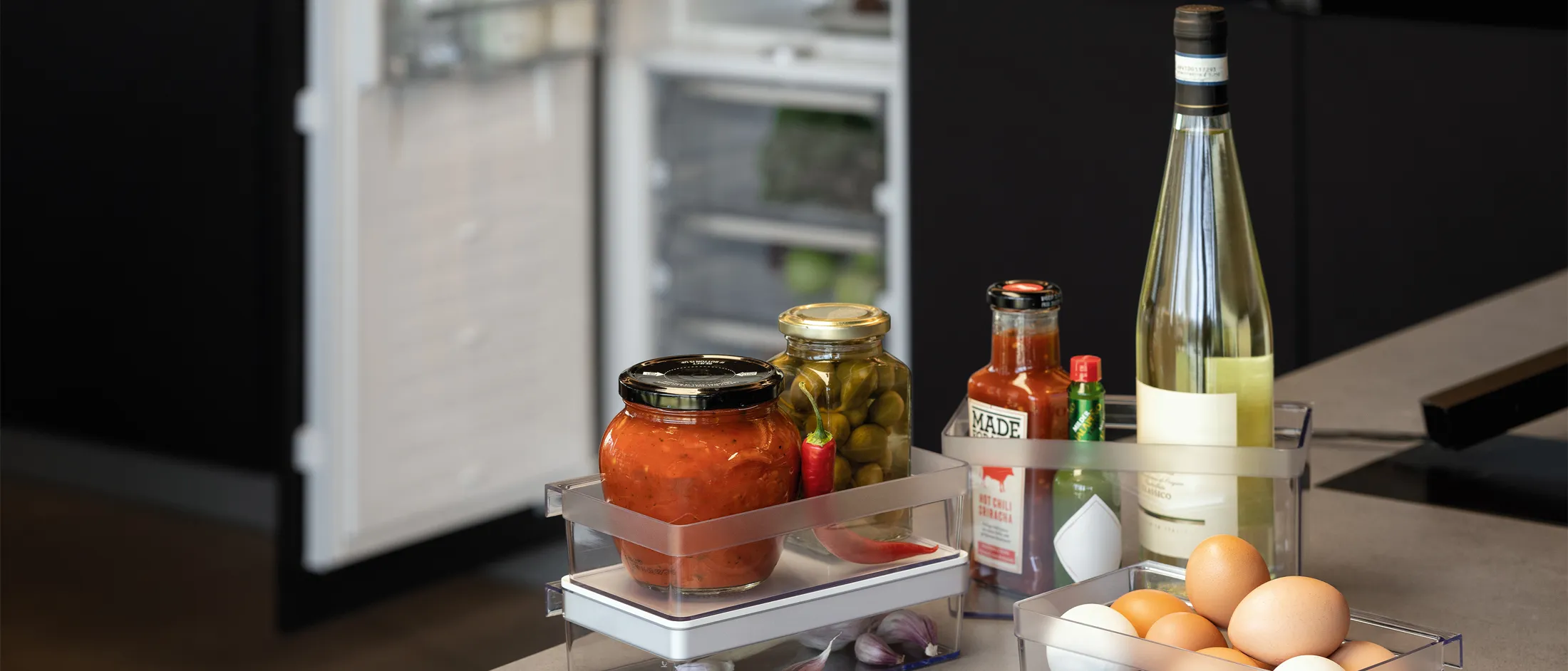 Condiments and eggs in multiple Flex Cooling storage boxes on the kitchen counter in the foreground with an open fridge and freezer in the background.