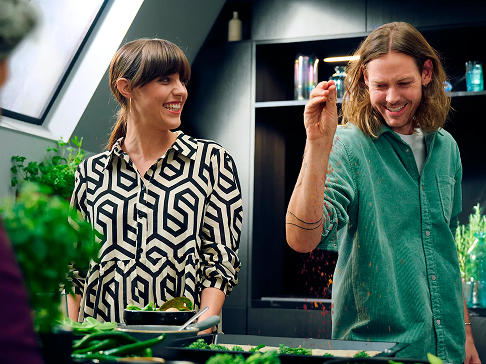 Man in black kitchen cooking with friends, throwing spice on dish on vented cooktop 