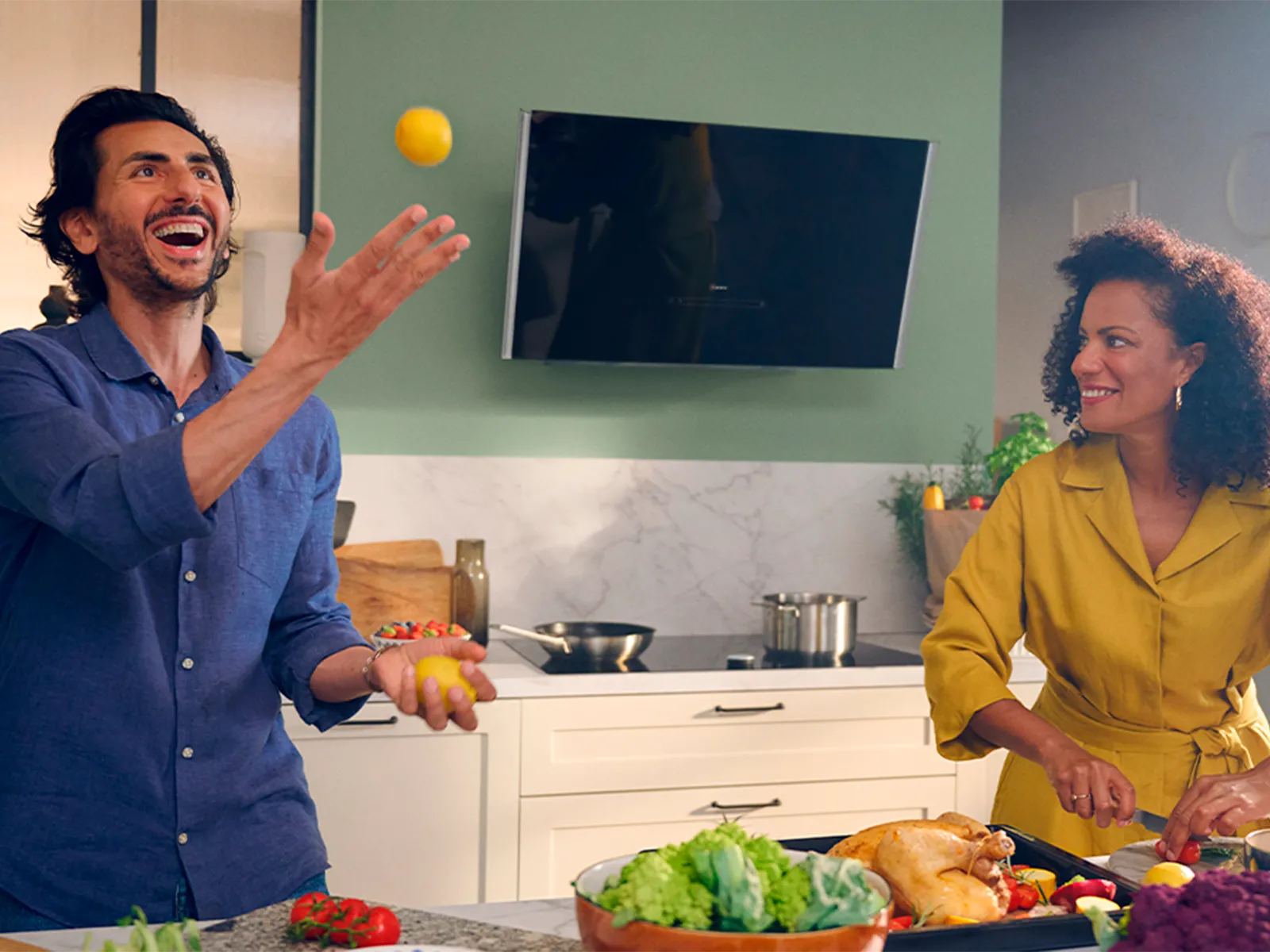 Man throwing lemon, woman laughing in front of inclined hood mounted on green wall 