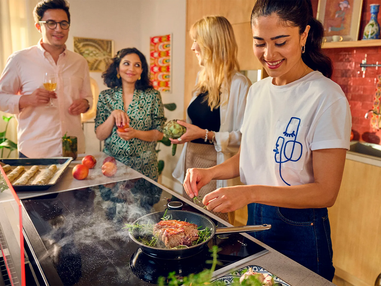 Four people in a kitchen, one cooking at the stove, others chatting with drinks.