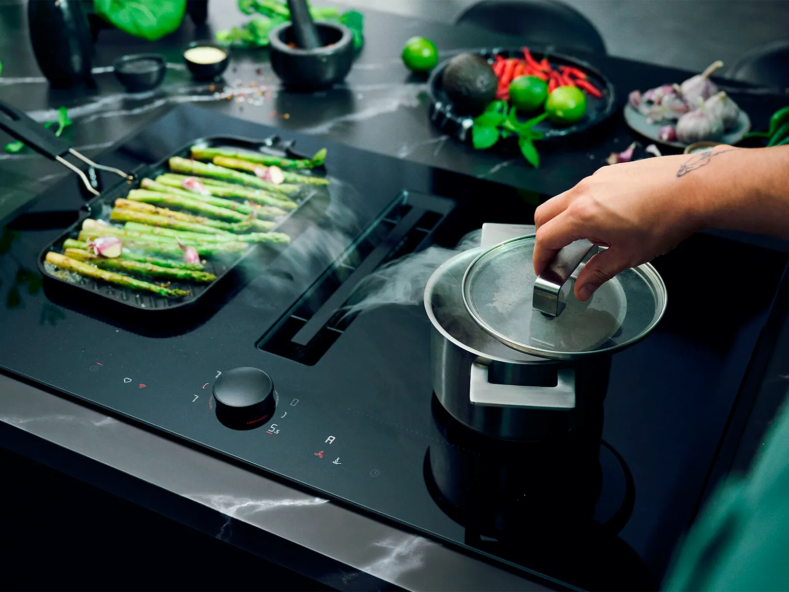 Hand lifting pot lid to release steam into vented cooktop, asparagus nearby 