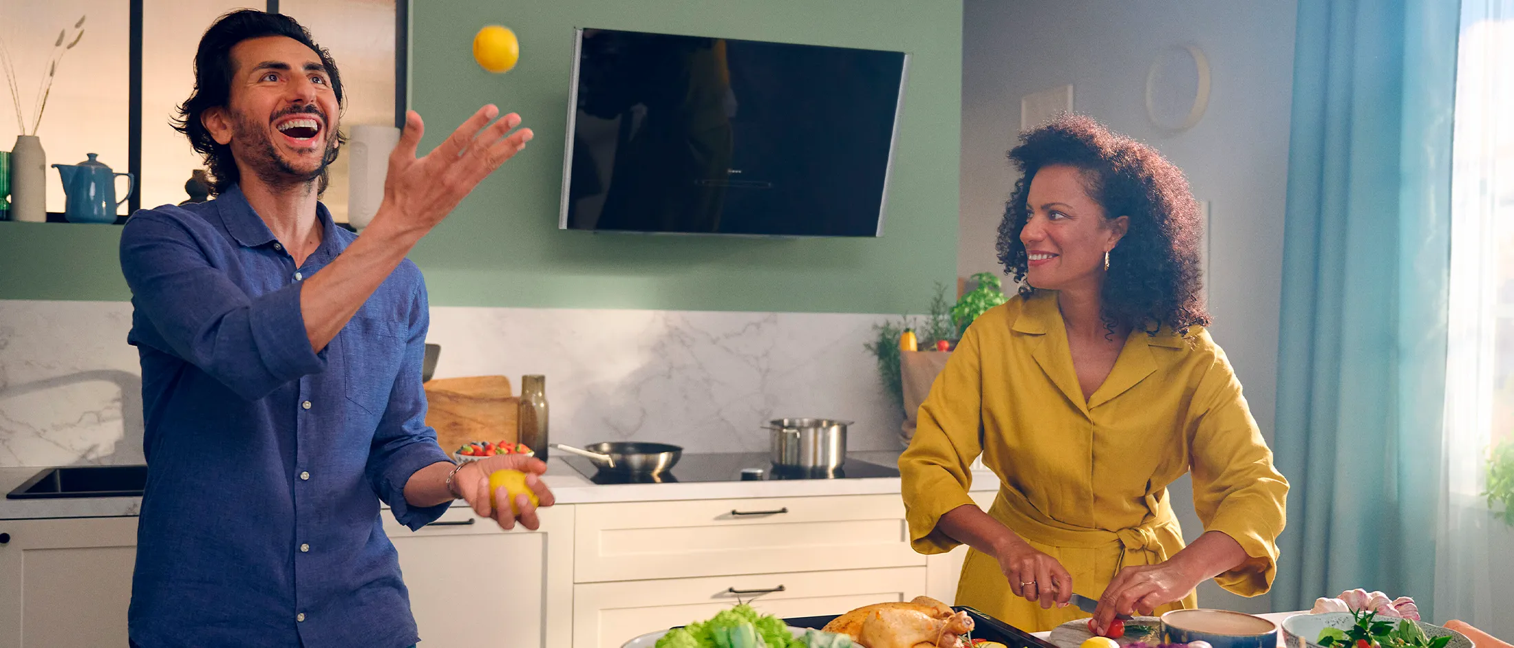 Man throwing lemon, woman laughing in front of inclined hood mounted on green wall 