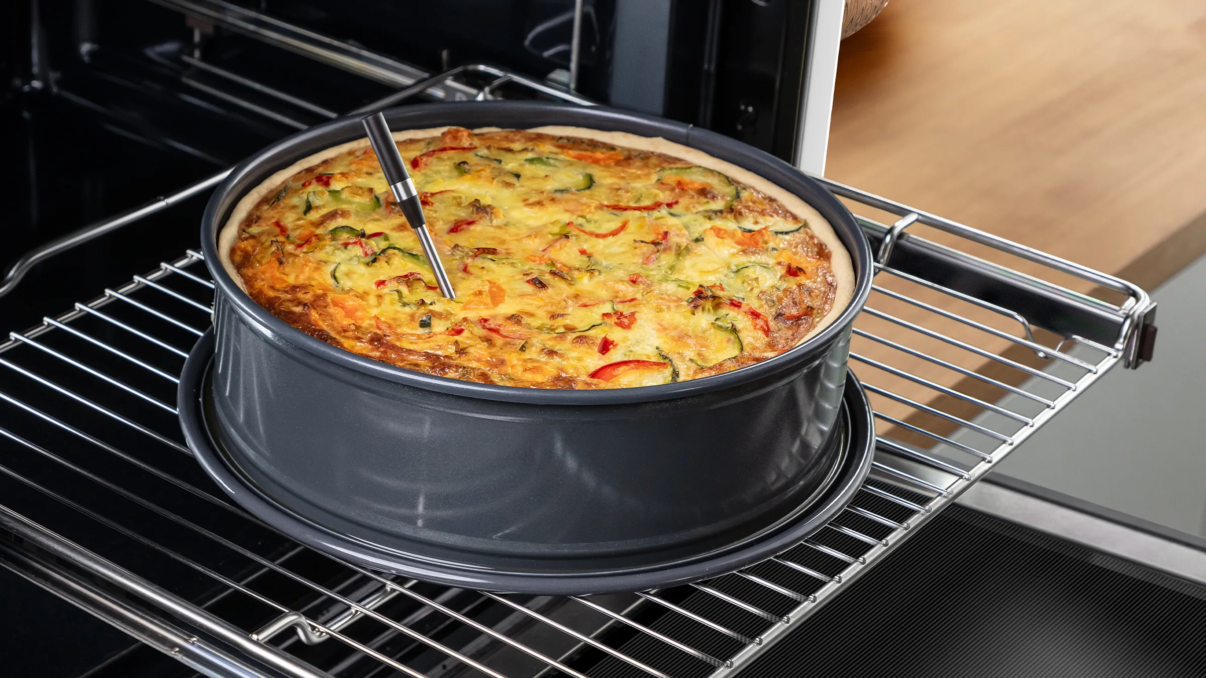 Frische Quiche in einer Springform auf dem ausgefahrenen Backofengitter eines Bosch-Backofens mit genutztem PerfectRoast Thermometer.