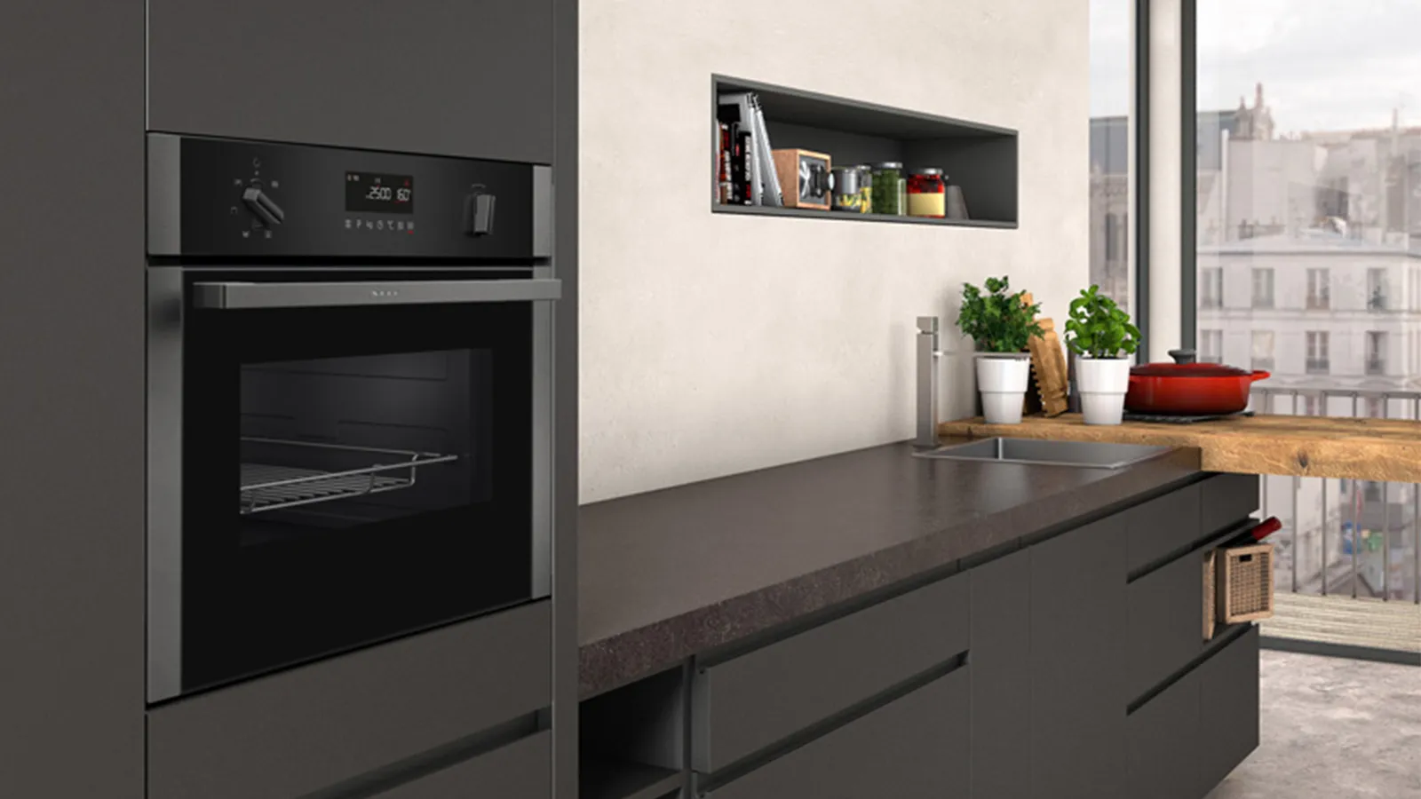 Contemporary kitchen with a built-in black wall oven at left, matte dark-gray lower cabinets and a long dark countertop holding potted herbs, a red bowl and a wooden cutting board; a recessed wall niche with jars and a large window overlooking city rooftops.
