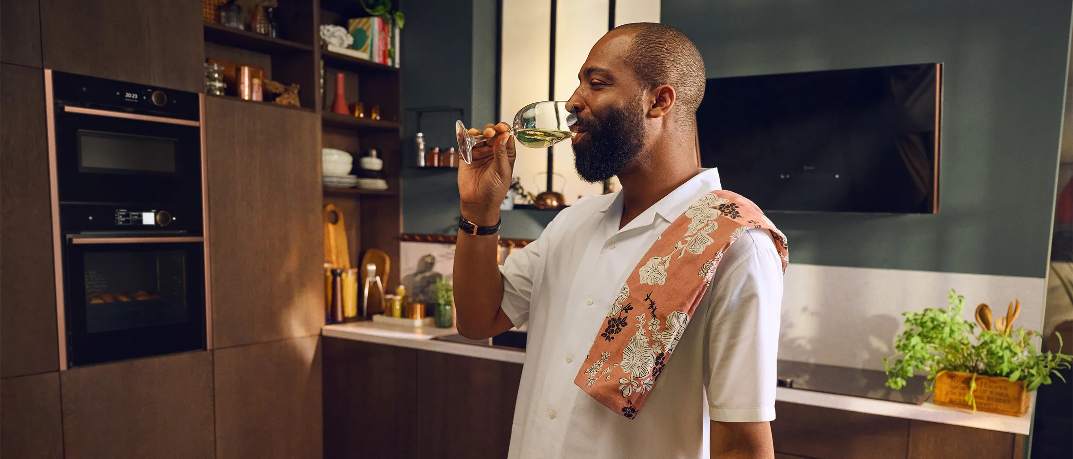 A person in a white shirt holds a glass of green liquid in a modern kitchen, draped with a floral patterned towel.