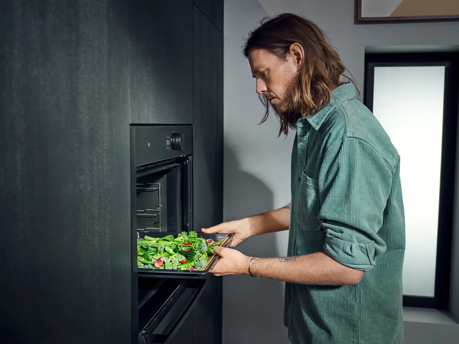 Man putting tray of green vegetables into single open oven in black kitchen
