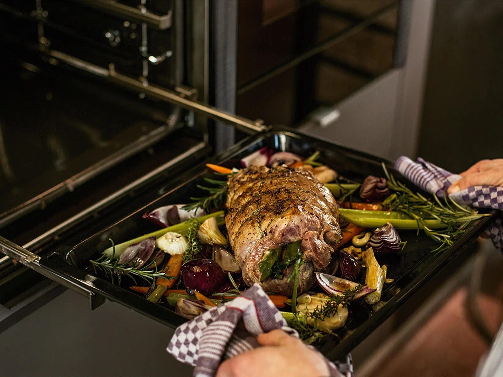 Oven tray filled with meat roast and vegetables being pulled out of oven on extendable rails