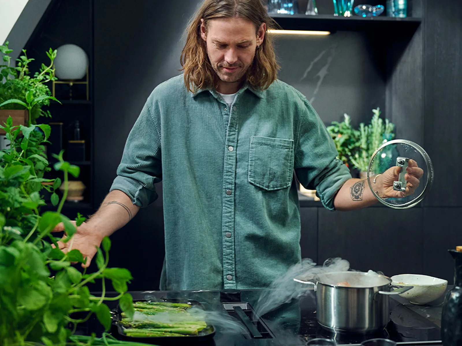 A man in a denim shirt cooking in a modern kitchen with pots on the venting hob.
