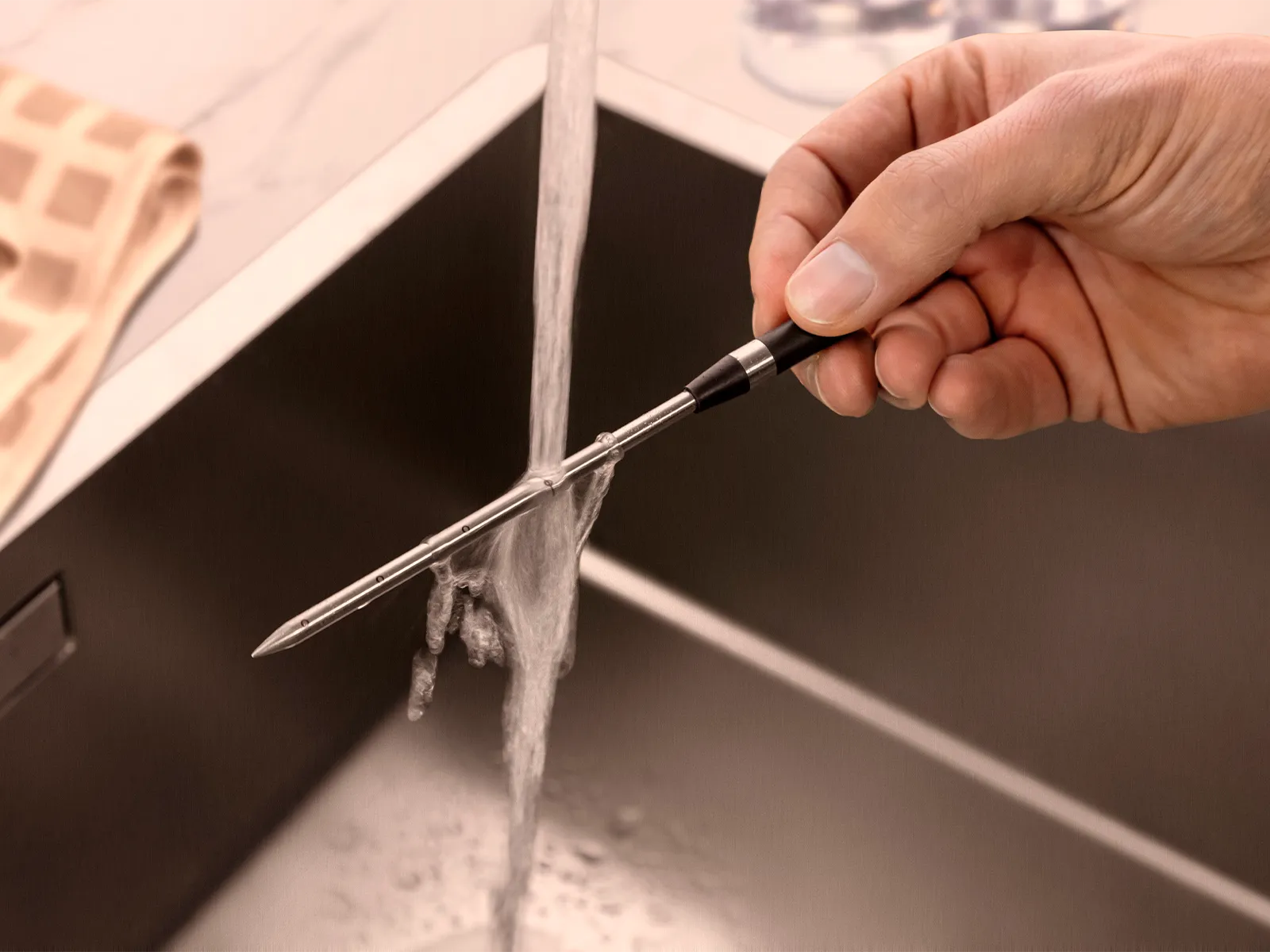 En hand håller en stektermometer under rinnande vatten i en diskho.