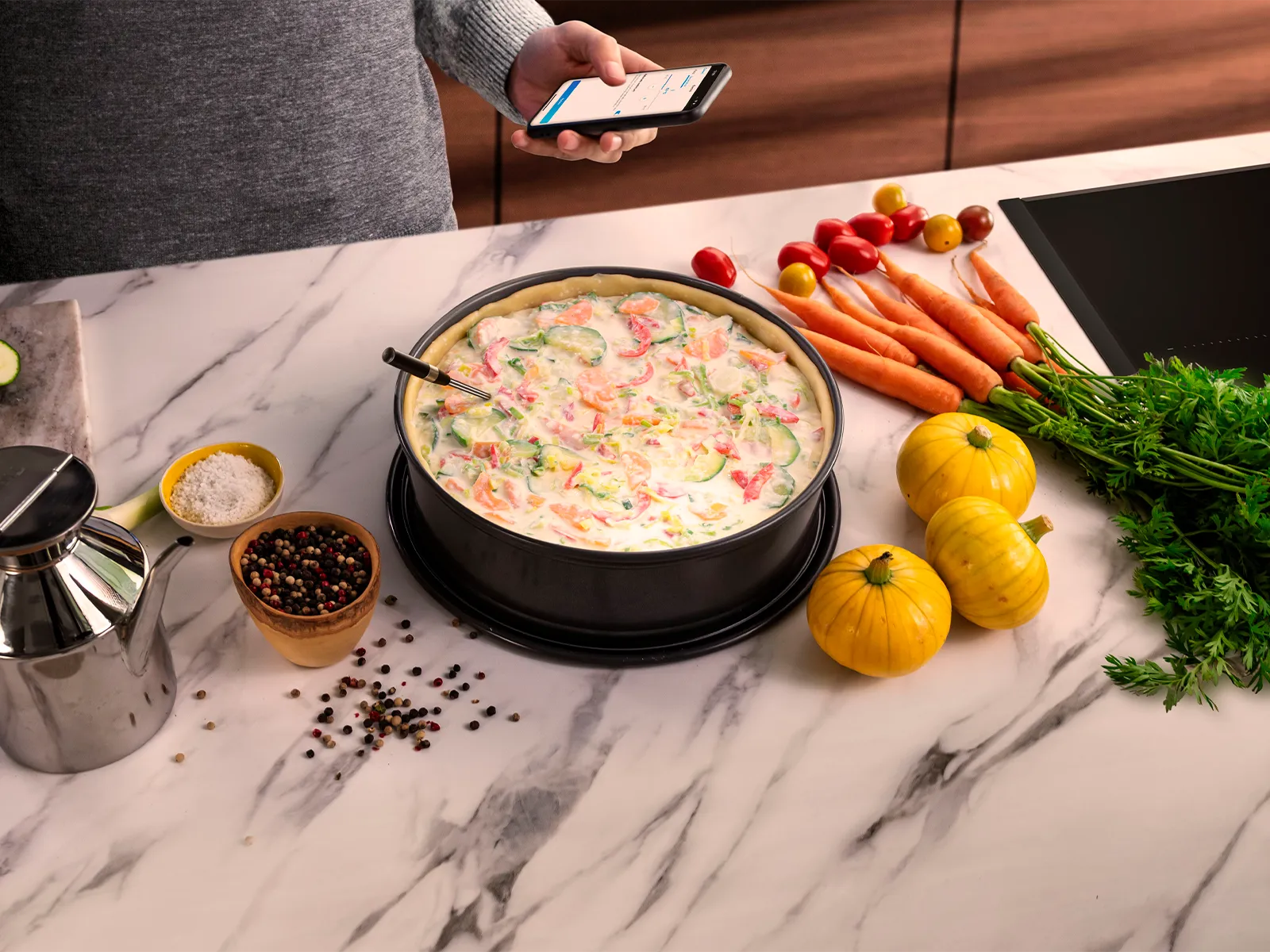 En paj i en rund svart form, omgiven av färggranna färska grönsaker, kryddor och en oljeflaska i rostfritt stål.