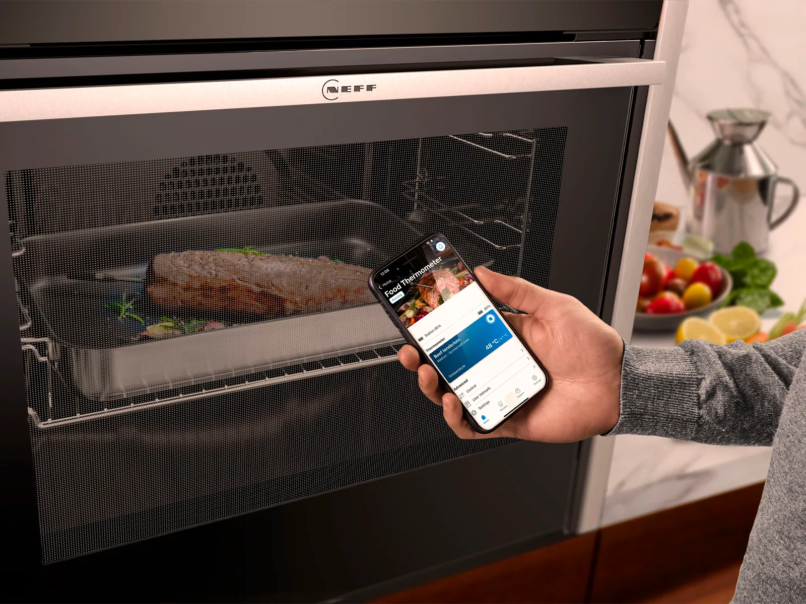 En hand håller en smarttelefon som visar tillagningsinformation, medan en laxfilé steks inuti en Neff-ugn.