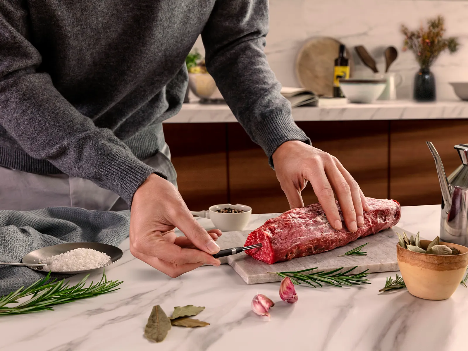 En person i en grå tröja sätter i en stektermometer i rått kött på en köksbänk i marmor.