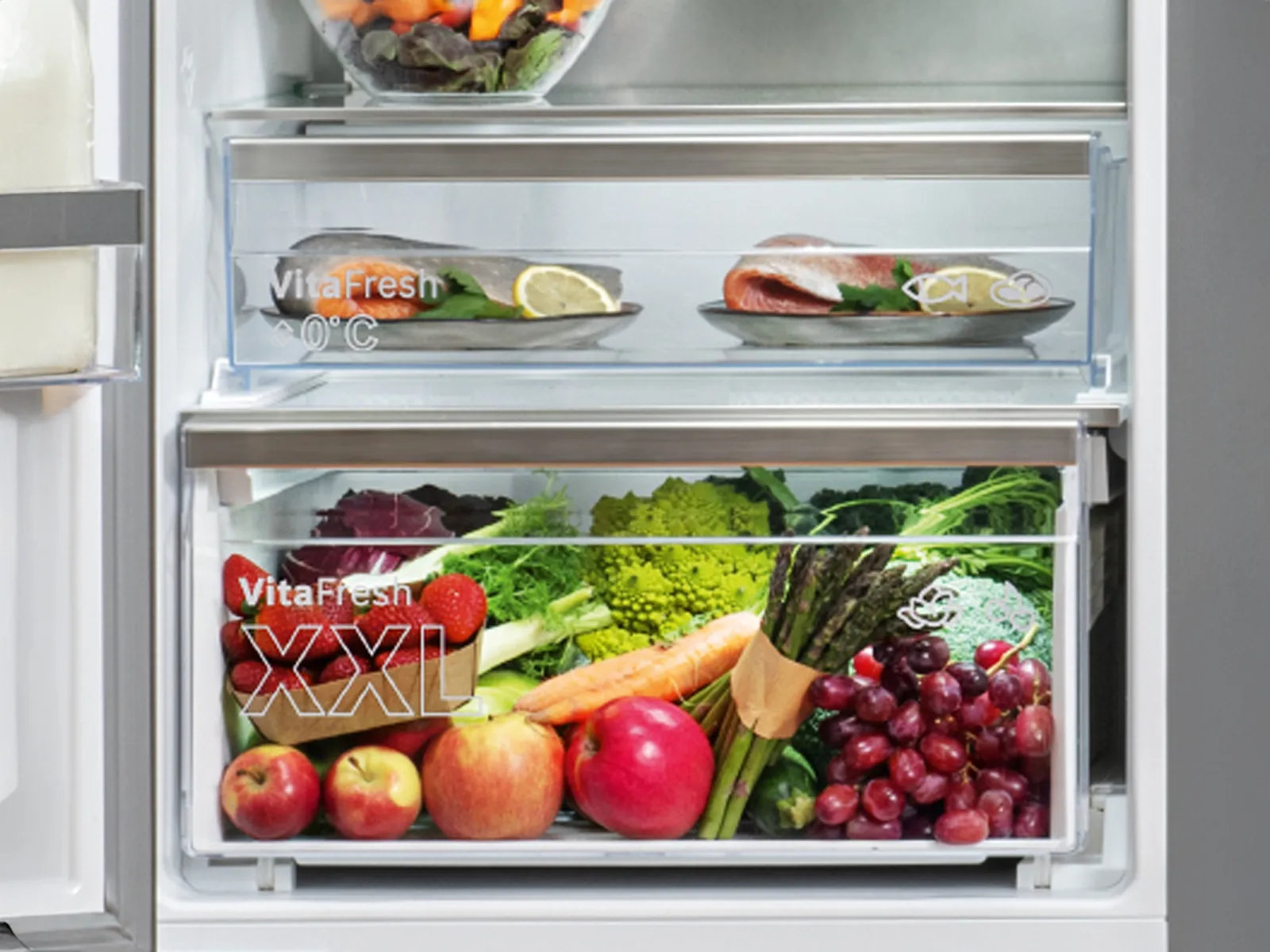 E Blick in en Kühlschrank, wo frisch Früchte und Gemüse in organisierten Schublade drinn sind, sowie Fisch uf e Teller darüber.