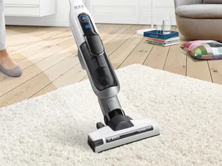 Person using a Bosch cordless vacuum cleaner on a white carpet in a bright living room.