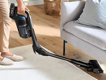 A person uses a cordless vacuum cleaner to clean a light carpet under a sofa in a cozy living room setting.