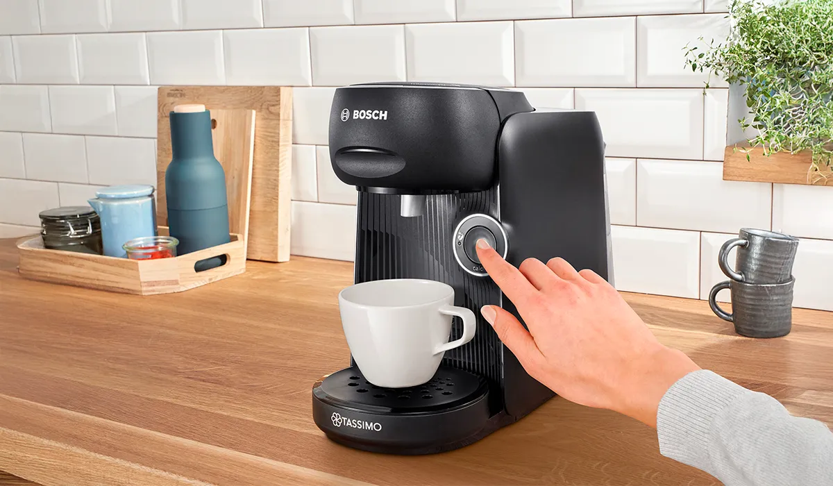 O persoană apasă pe butonul de start al mașinii de cafea prietenoase TASSIMO FINESSE.