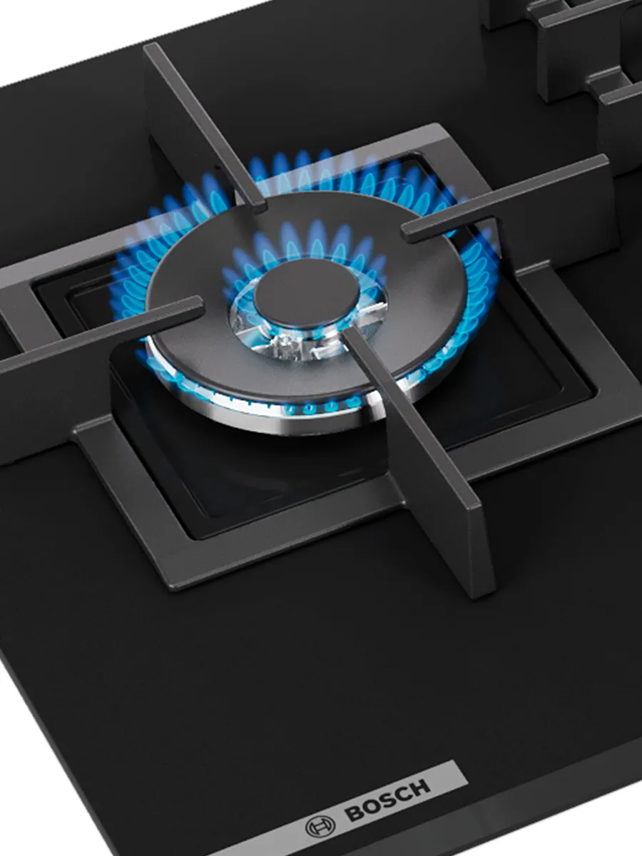 A modern gas stovetop with a lit burner producing blue flames, next to an olive oil bottle and wooden spoon.