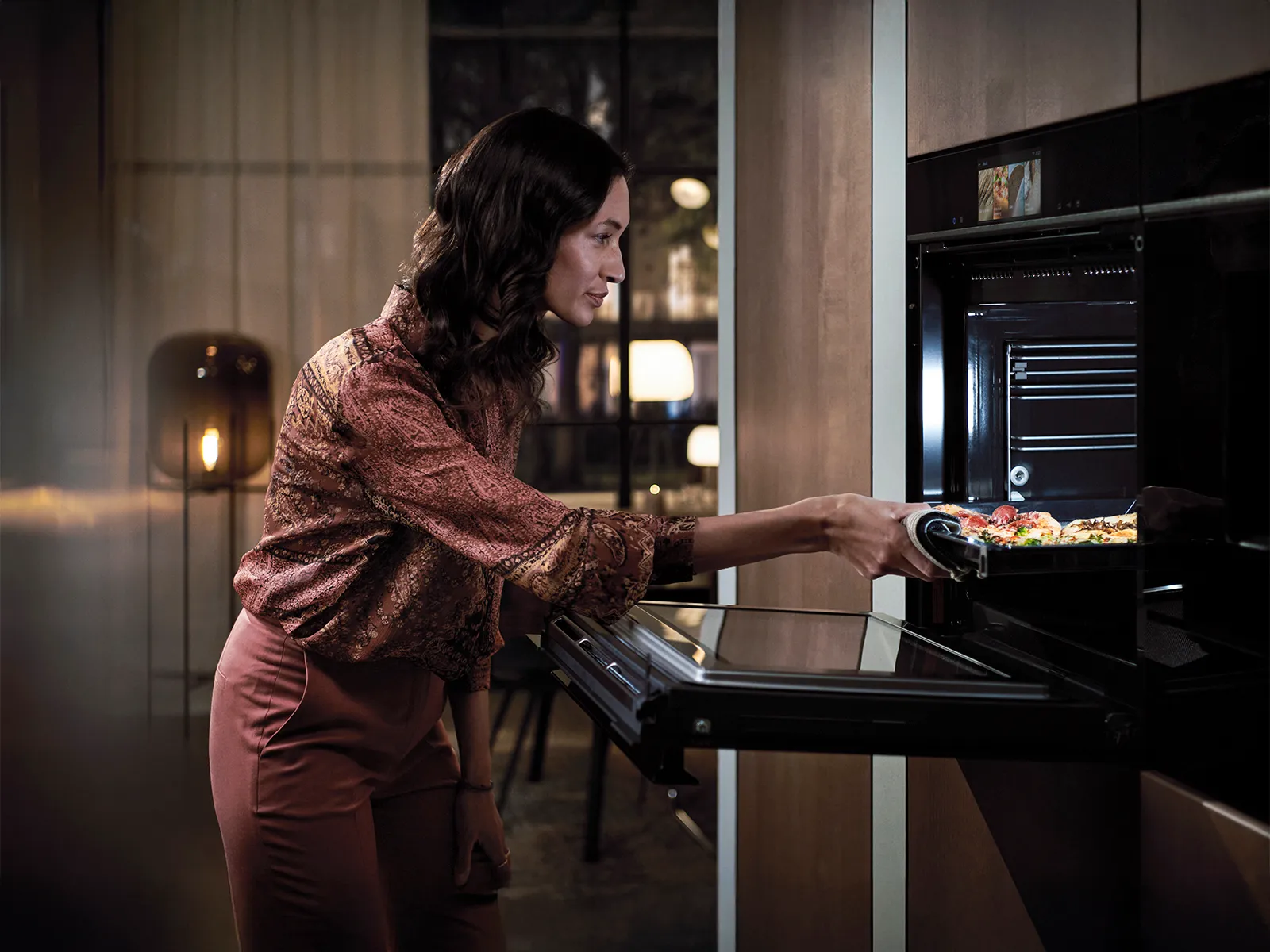En kvinne i stilige klær strekker seg inn i en moderne ovn for å hente et brett med fargerike grønnsaker i et varmt belyst kjøkken.