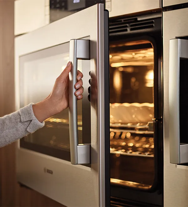 Woman opening Bosch SideOpening Wall Oven with dark brown cabinets in the background and a tray inside the oven