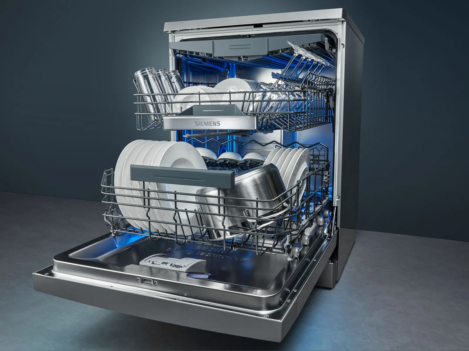 An open Siemens dishwasher filled with clean dishes, glasses, and pots, illuminated by blue lighting against a dark background.
