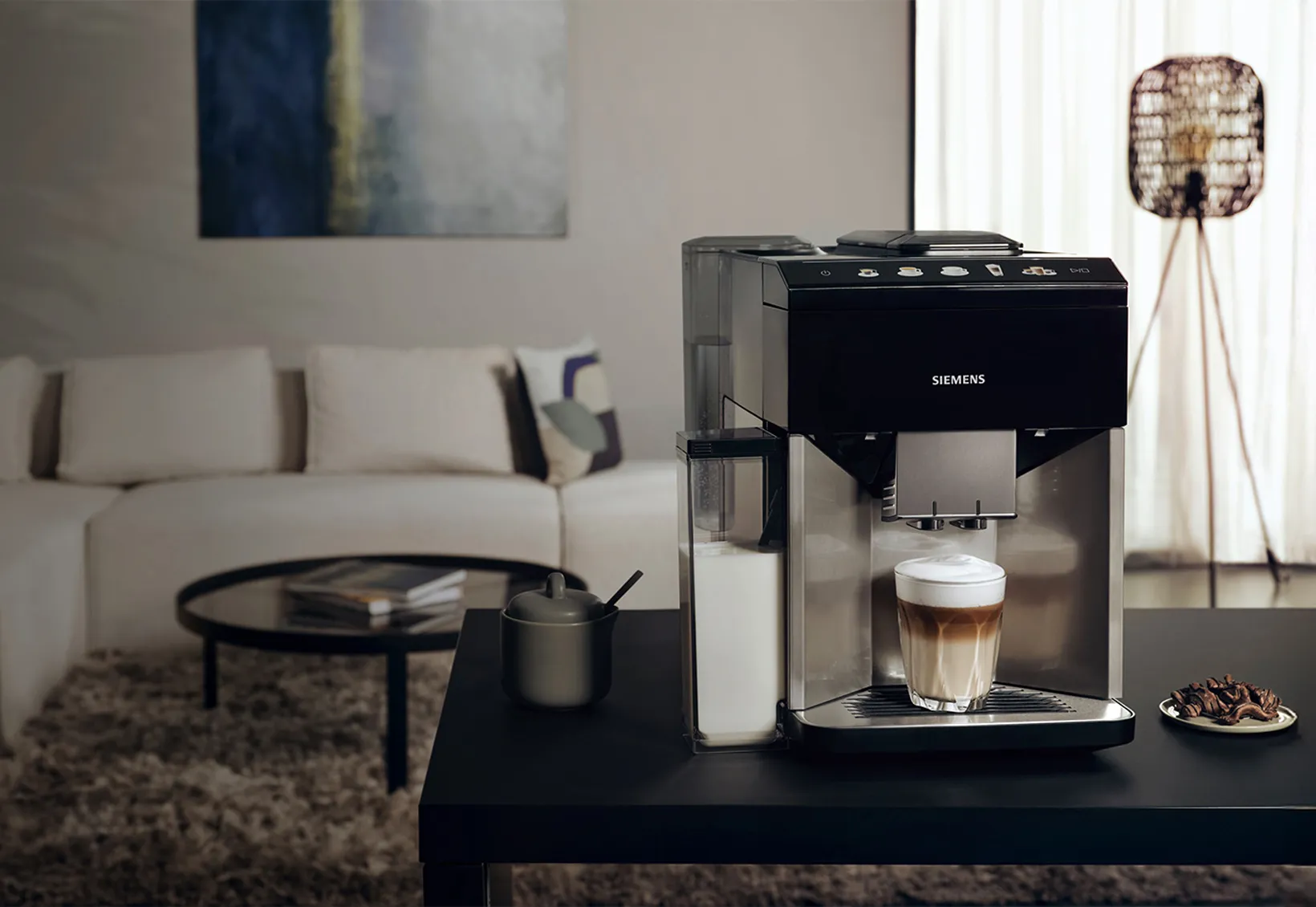 I forgrunden står EQ500 med en cappuccino under udskænkningen på en køkkenø; i baggrunden er en beige sofa.