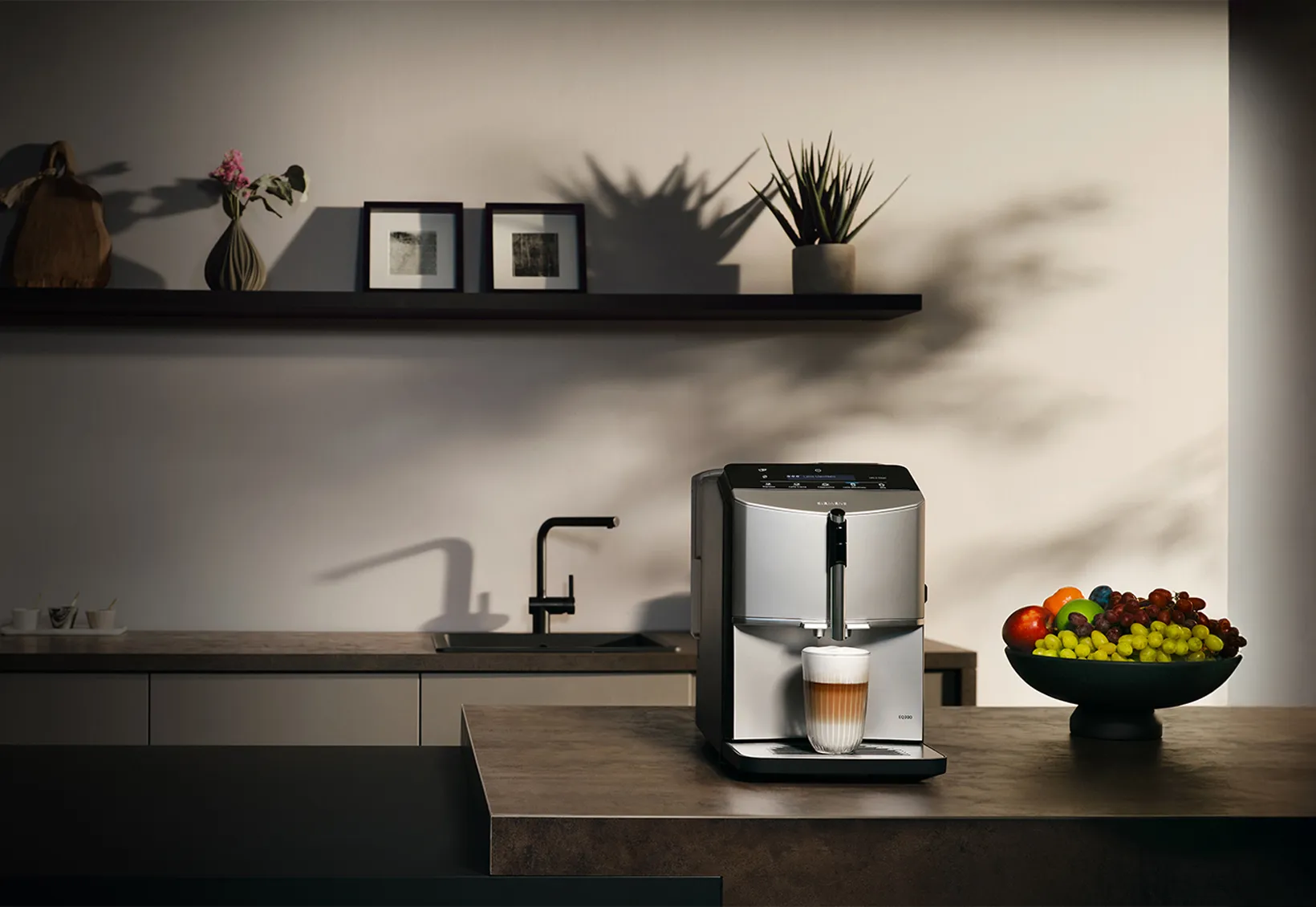 Køkken med EQ300 fuldautomatisk espresso-/kaffemaskine på en køkkenø, en kop latte macchiato står under udskænkningen. 