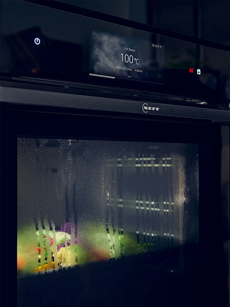 A black NEFF steam oven is shown seamlessly integrated into off-white kitchen cabinets. The oven door is fogged up with condensation from the steam, and the digital display shows it is operating in "Full Steam" mode at 100°C.
