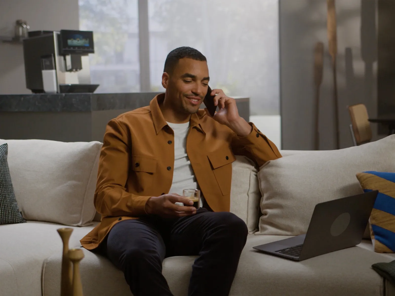 Un bărbat într-o jachetă bej stă pe o canapea cu un laptop și un smartphone.