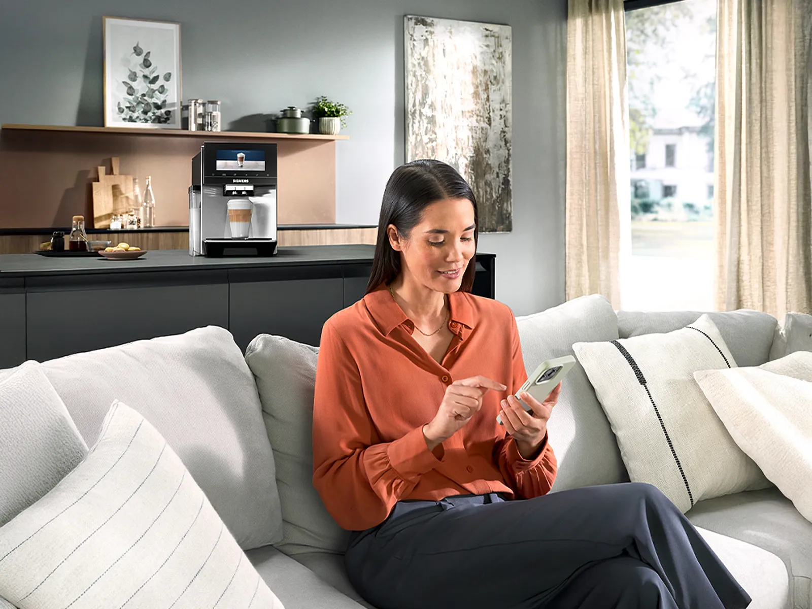 O femeie stă pe canapea într-un living, ținând un telefon în mână, cu o bucătărie modernă în fundal.