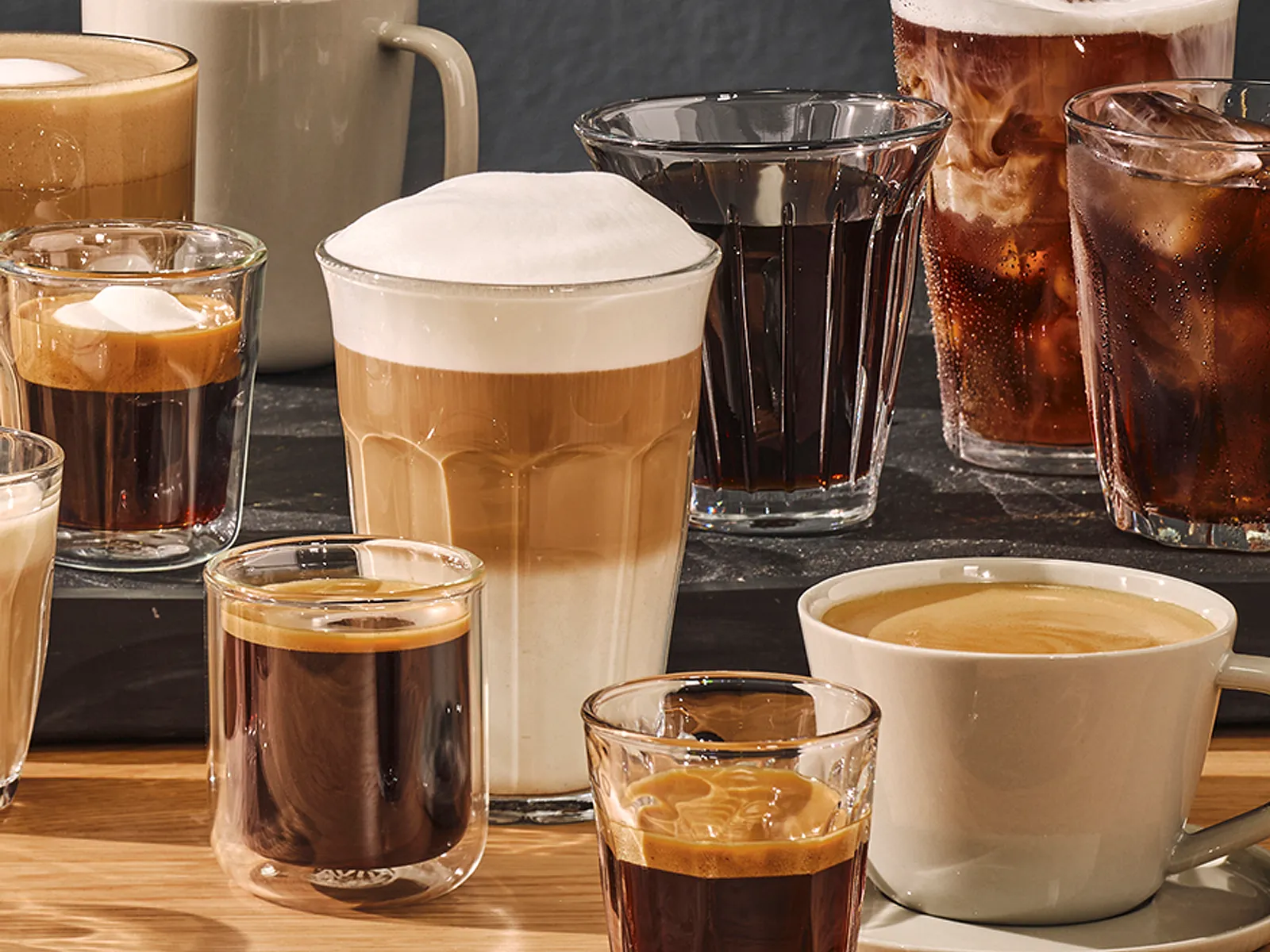 O varietate de băuturi pe bază de cafea așezate pe un blat de lemn, inclusiv un latte, espresso, cappuccino, flat white, Americano și cafea cold brew.