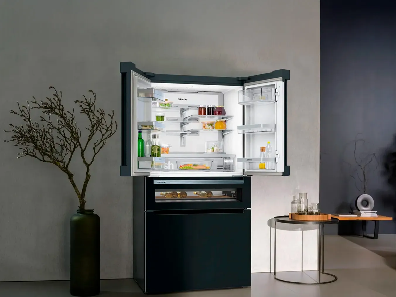 Moderno frigorifero nero con porte-finestre aperte, che espone vari cibi e bevande all'interno, accanto a un alto vaso e un piccolo tavolo rotondo con bicchieri.