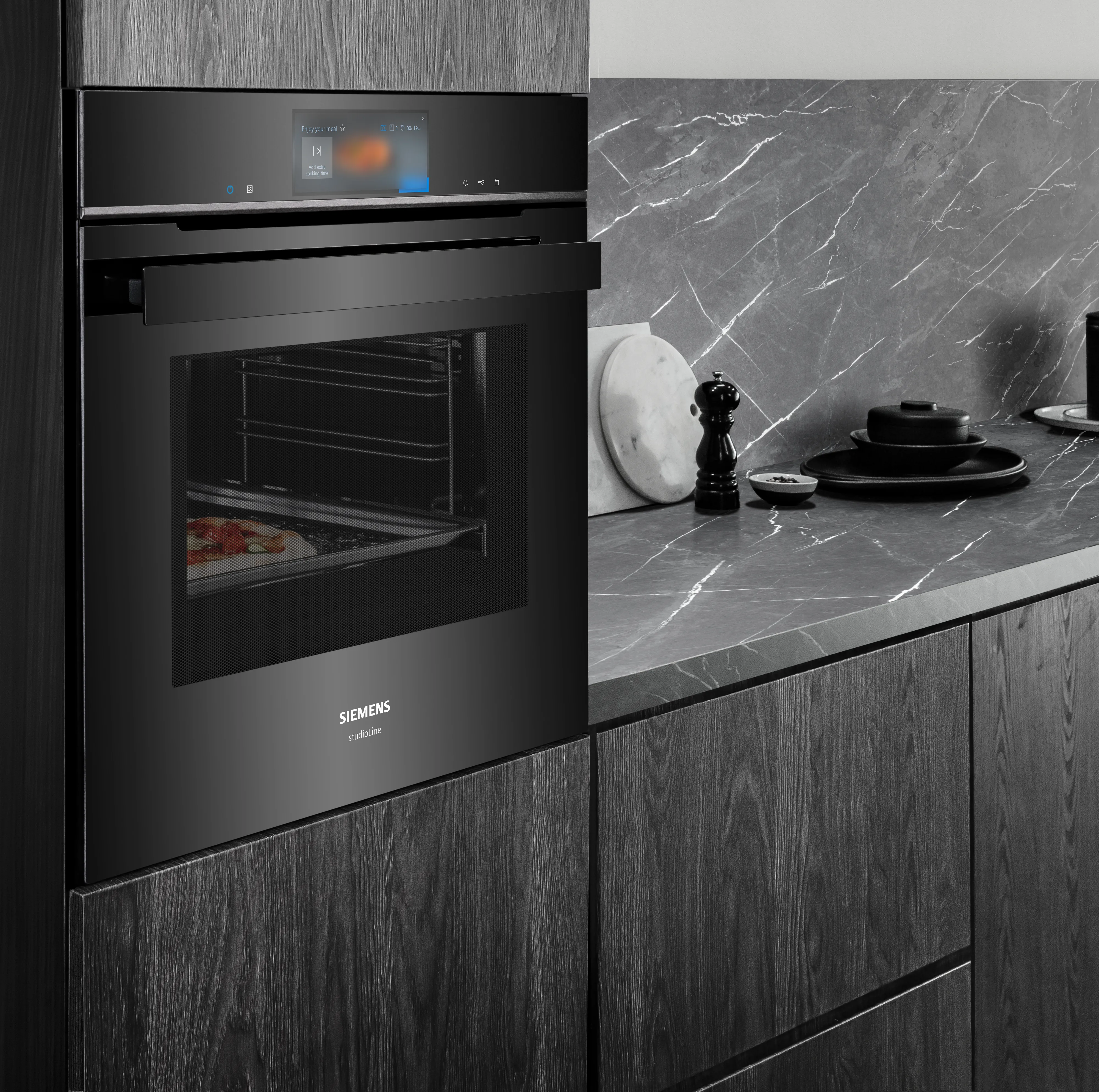 A close-up of a black Siemens iQ700 built-in oven, integrated into dark wood-grain cabinetry. A pizza is visible cooking inside the illuminated oven, and the digital display is active.