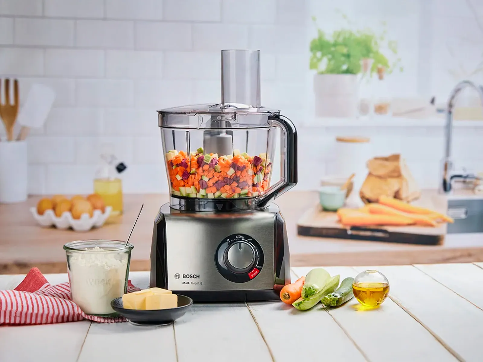 Robot de cuisine Bosch avec des légumes hachés, du fromage, de la farine et des ingrédients frais sur un plan de travail.