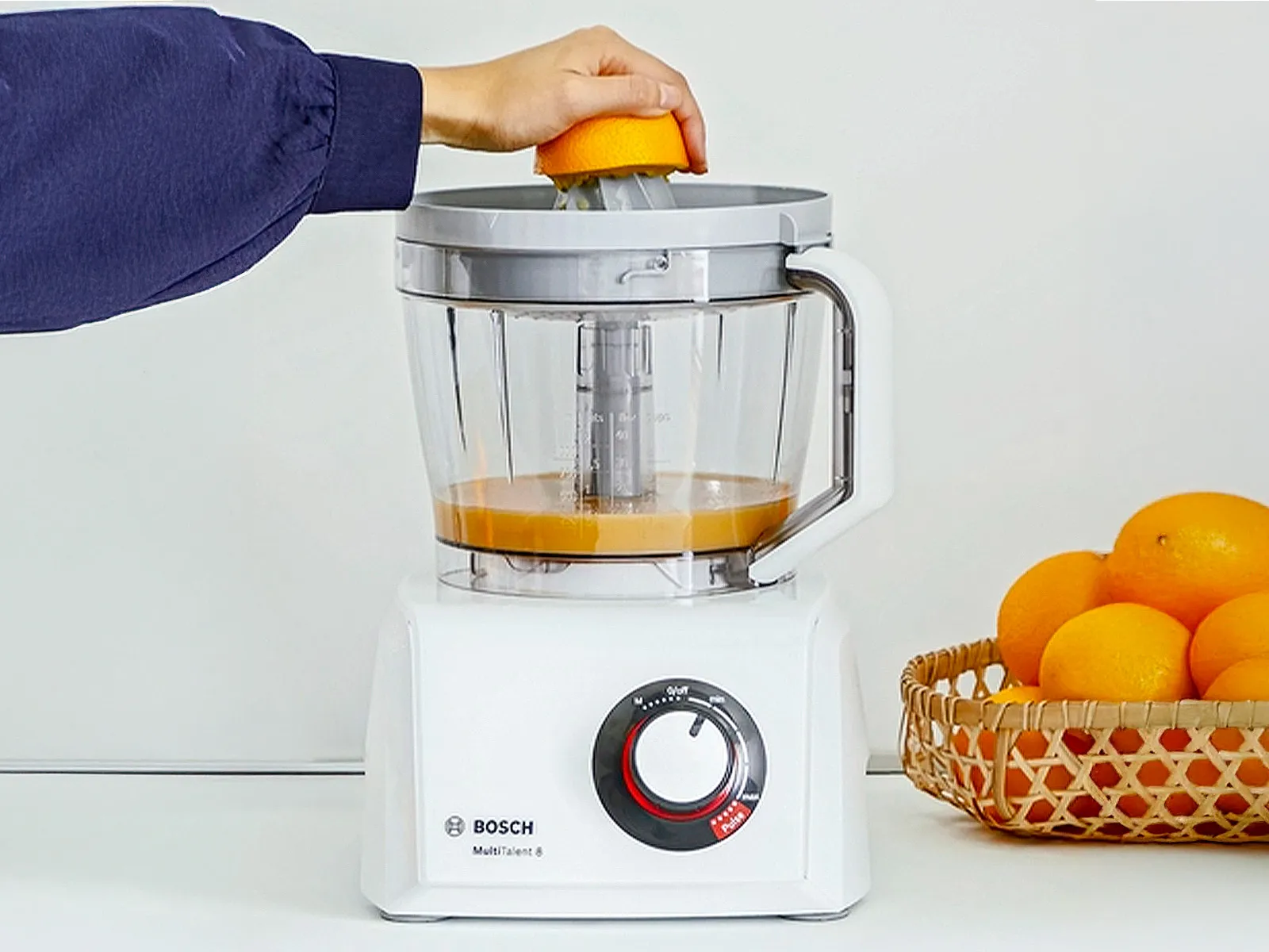 Une personne presse une orange avec un robot de cuisine Bosch MultiTalent 8, un panier d’oranges à côté.