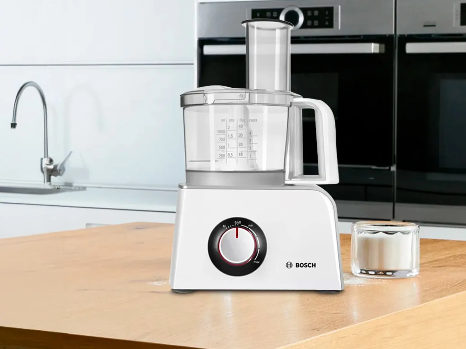 A Bosch food processor in white on a wooden countertop and a glass of sugar beside it.