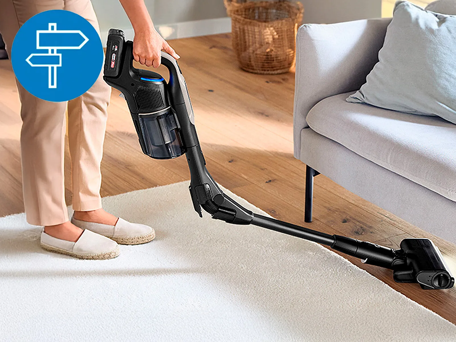 Osoba používající bezdrátový vysavač na bílé koberci, s ikonkou směrového značení v rohu.