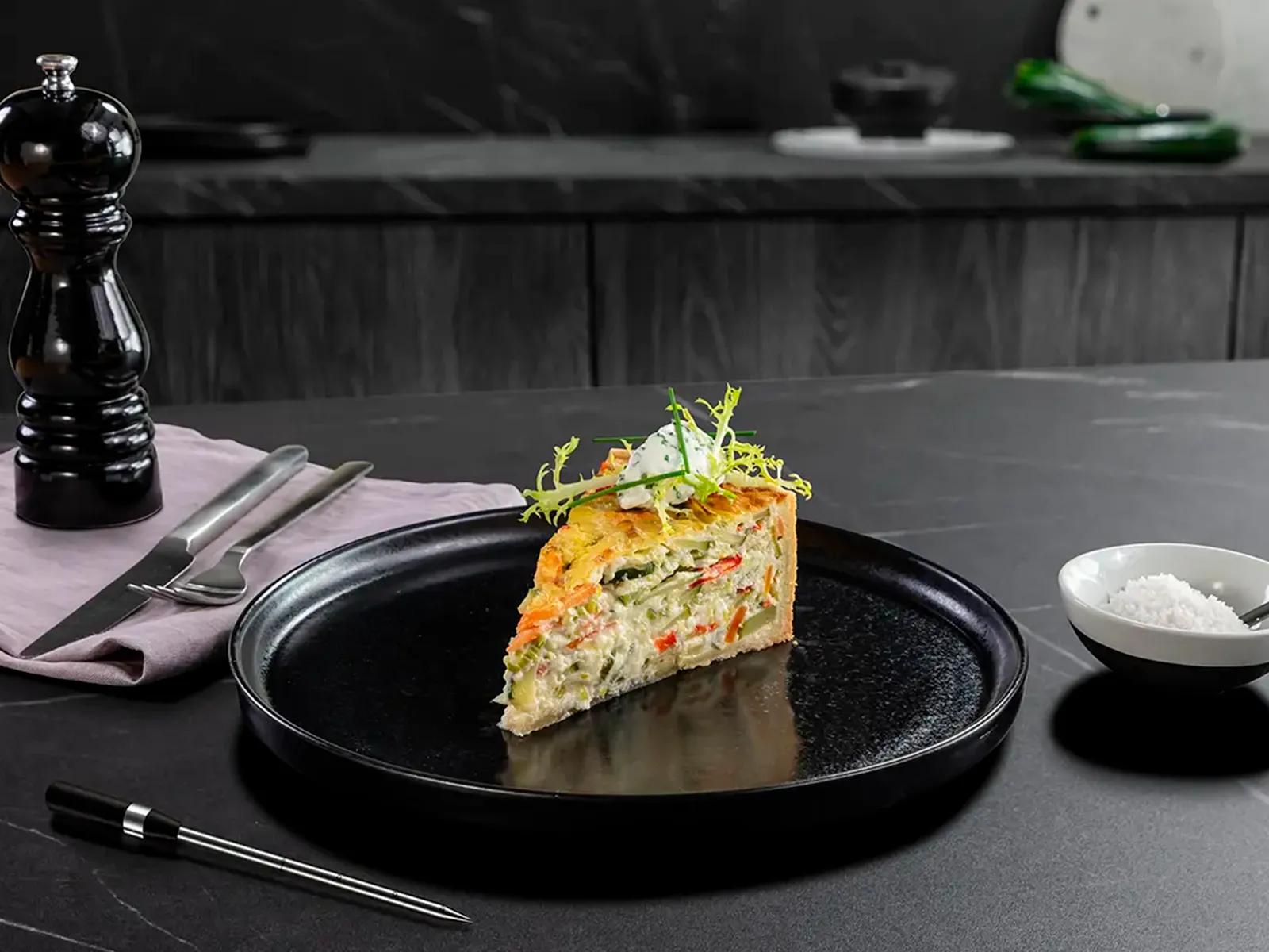 Ein Stück Gemüsequiche auf einem schwarzen Teller auf einer dunkelgrauen Arbeitsplatte neben einer Pfeffermühle.
