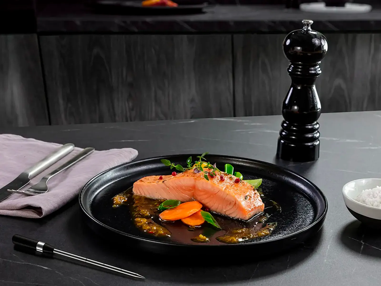 Ein gegartes Lachsfilet mit Sauce auf einem schwarzen Teller, auf einer grauen Arbeitsplatte neben einer Pfeffermühle.
