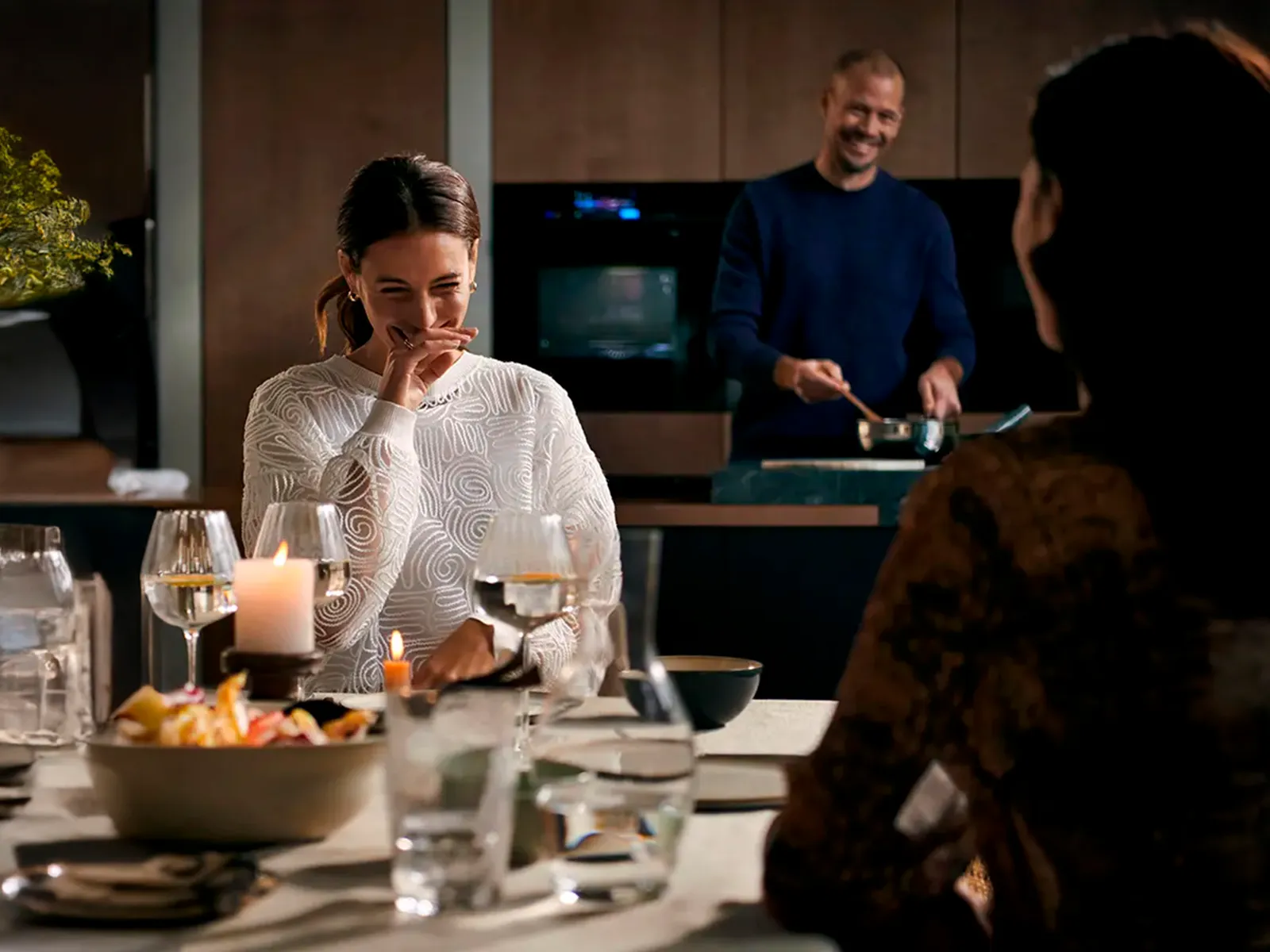 Zwei Frauen sitzen am Esstisch, während im Hintergrund ein Mann in der Küche Essen anrichtet.