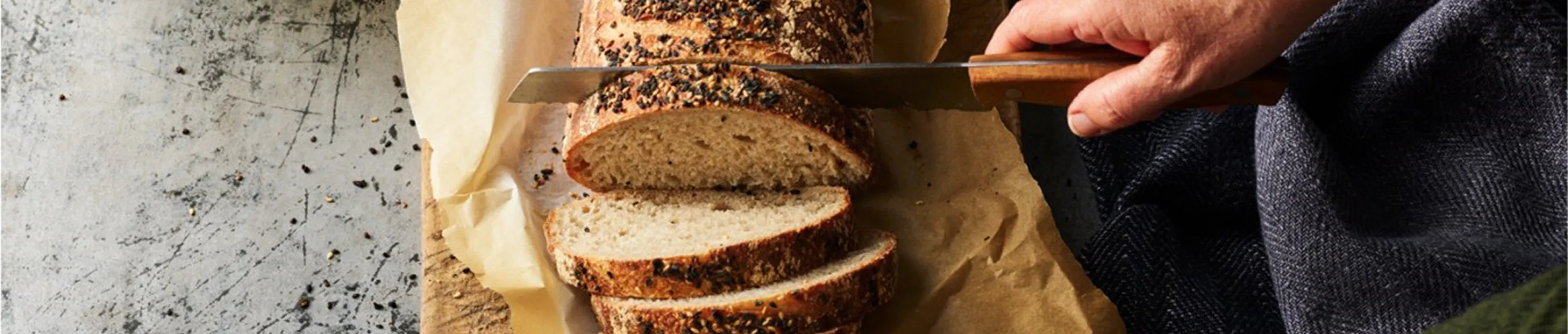 A hand slicing seeded bread on parchment paper