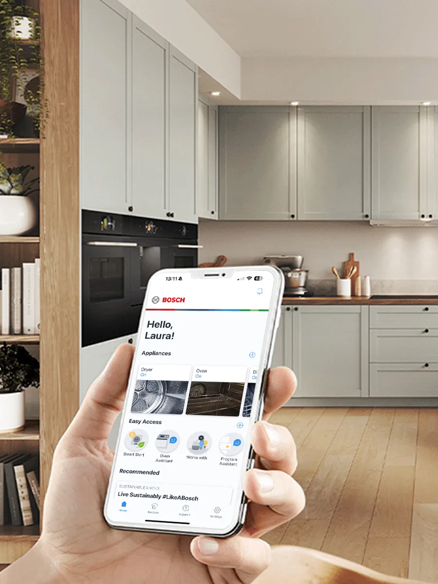 Eine Hand hält ein Smartphone mit der Bosch Home Connect App-Startseite in einer modernen, hellgrauen Küche mit Einbaugeräten.