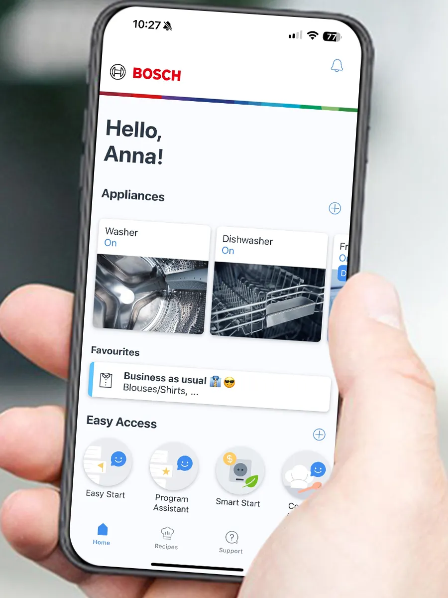 Eine Hand hält ein Smartphone mit der personalisierten Bosch Home Connect Startseite, die eine eingeschaltete Waschmaschine und einen Geschirrspüler anzeigt.