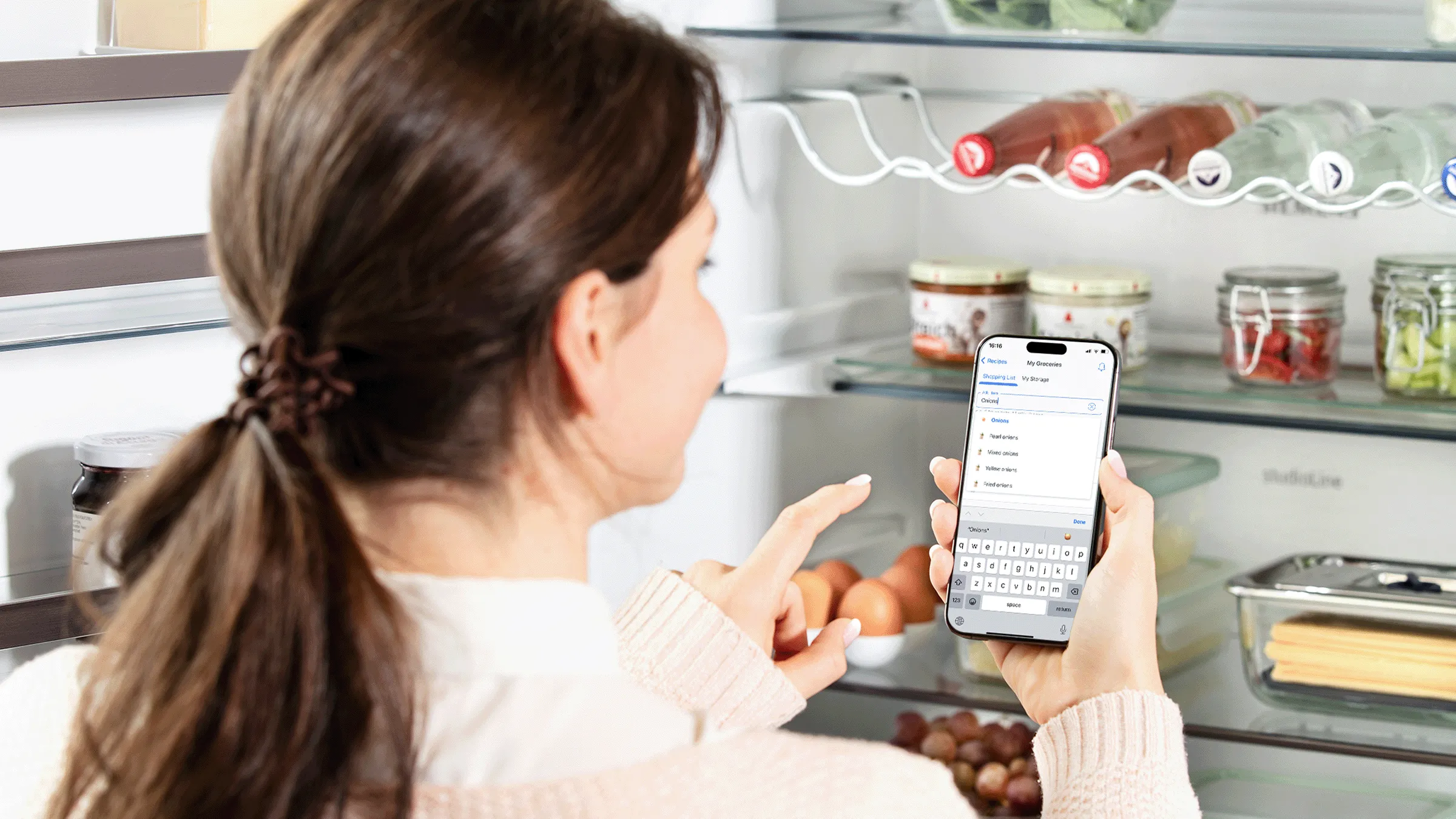 Eine Frau blickt in ihren geöffneten Kühlschrank und nutzt dabei die Home Connect App auf ihrem Smartphone.